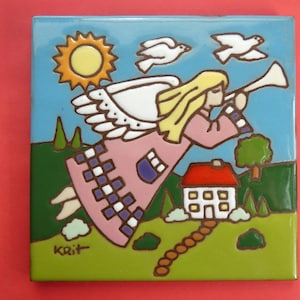 May include: A square ceramic tile featuring a colorful illustration of a blonde angel in a pink robe playing a trumpet. The scene includes a yellow sun, two white birds, a house with a red roof, and green hills against a blue sky. The artist signature KRIT is visible in the bottom left corner.