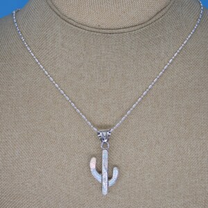 May include: A silver necklace with a cactus pendant. The pendant is white with iridescent accents, and the chain is a delicate silver. The cactus design is a stylized representation of the desert plant.