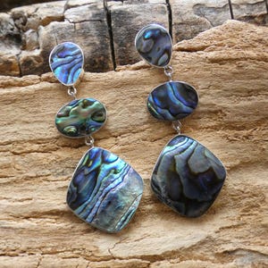 Può includere: Un paio di orecchini d'argento con tre gocce di abalone iridescente. La goccia superiore è rotonda, la goccia centrale è ovale e la goccia inferiore ha una forma di diamante.