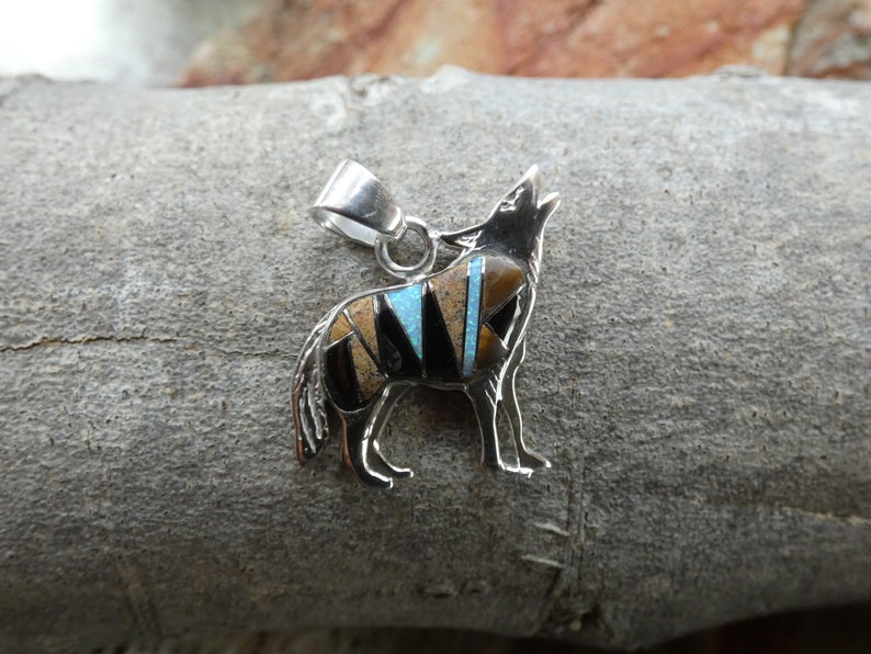 May include: Sterling silver pendant featuring a howling wolf design. The wolf is inlaid with turquoise, black onyx, and tiger eye stones.