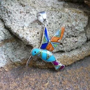 Op de afbeelding: Een zilveren kolibrie-hanger met turquoise, oranje en paarse inleg. De hanger is gemaakt van sterling zilver en is een prachtig voorbeeld van Native American sieraden.