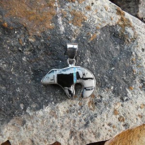 May include: A silver pendant shaped like a bear, featuring inlaid segments of white and black stone with a thin vertical stripe of iridescent blue opal. The pendant includes a silver bail at the top for attachment to a necklace chain.