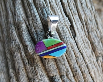 May include: A silver pendant with an oval-shaped inlay of colorful stones. The stones are arranged in a geometric pattern and include shades of green, purple, red, blue, and yellow.
