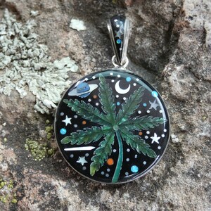 Puede incluir: Un colgante de plata con fondo negro y un diseño de hoja de marihuana verde. El colgante está decorado con pequeñas estrellas blancas, una luna creciente y un planeta azul con un anillo alrededor.