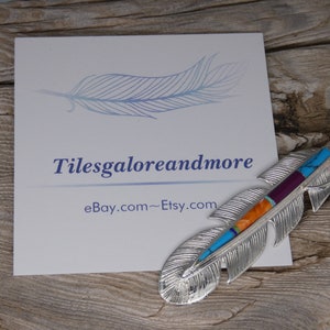 May include: A silver feather pendant with turquoise, orange, purple, and red inlay. The pendant is on a wooden surface with a business card that says "Tilesgaloreandmore eBay.com~Etsy.com".