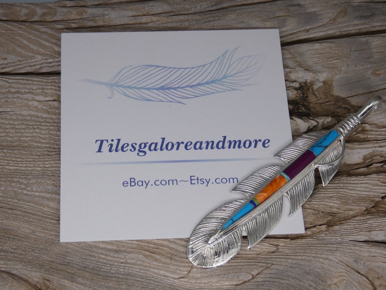 May include: A silver feather pendant with turquoise, orange, purple, and red inlay. The pendant is on a wooden surface with a business card that says "Tilesgaloreandmore eBay.com~Etsy.com".
