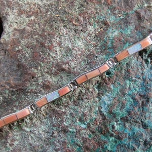 May include: A silver bracelet with rectangular coral and turquoise-colored stone inlays. The bracelet is set against a textured, multi-colored stone background. The bracelet's design features alternating coral and turquoise segments.