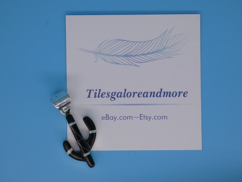 May include: A black cactus pendant with turquoise accents. The pendant is on a white card with the text "Tilesgaloreandmore" and "eBay.com~Etsy.com" printed on it. The background is a solid light blue.