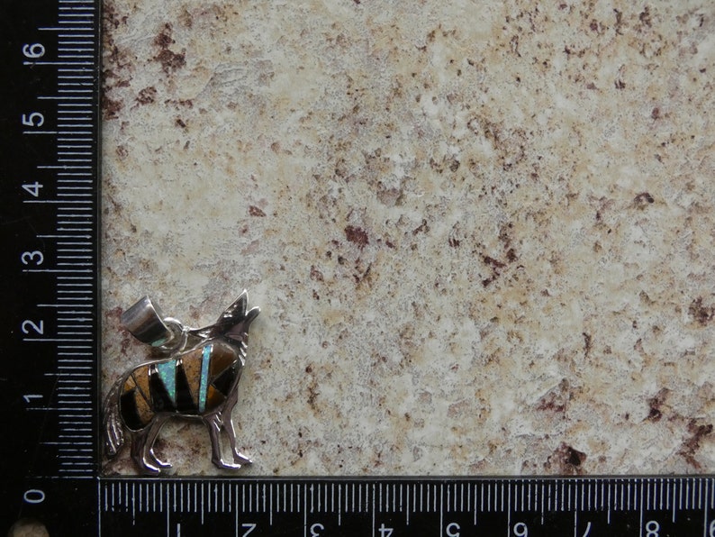 May include: A silver pendant featuring a howling wolf with inlaid turquoise, black, and brown stones. The pendant is approximately 2 inches tall.