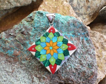 May include: A square pendant with a colorful flower design made of inlaid stones. The pendant is white with red, green, blue, and orange stones. The pendant has a silver bail.