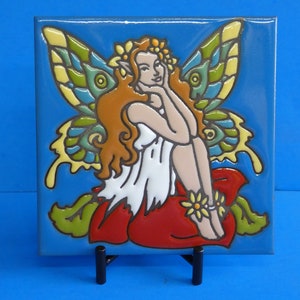 May include: A ceramic tile with a hand-painted image of a fairy with wings, sitting on a red flower. The fairy has long red hair, a white dress, and a flower crown. The background is blue.