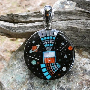 May include: A round silver pendant with a black background and colorful inlay depicting a stylized galaxy with stars, planets, and a comet. The pendant is made of sterling silver and features a variety of gemstones, including turquoise, coral, and mother-of-pearl.