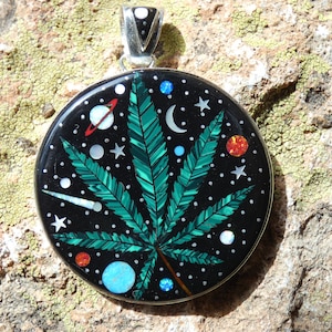 Puede incluir: Un colgante redondo de plata con un diseño de hoja de marihuana verde sobre un fondo negro con estrellas blancas, una luna creciente y planetas. El colgante está hecho de piedra incrustada y es una pieza de joyería única.