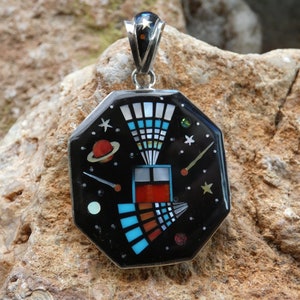 Puede incluir: Un colgante de plata con fondo negro e incrustaciones de colores que representan una escena espacial estilizada con un planeta, estrellas y un cometa. El colgante tiene forma octogonal y tiene una anilla de plata.