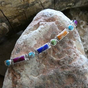 May include: A silver bracelet with rectangular and circular gemstone accents in various colors, including blue, orange, green, and purple. The bracelet is displayed on a textured rock surface, showcasing the jewelry's design and craftsmanship.