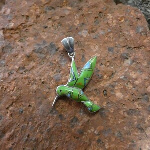 May include: A green and blue hummingbird pendant made of sterling silver. The hummingbird is in flight with its wings spread out. The pendant is a beautiful piece of jewelry that would make a great gift.