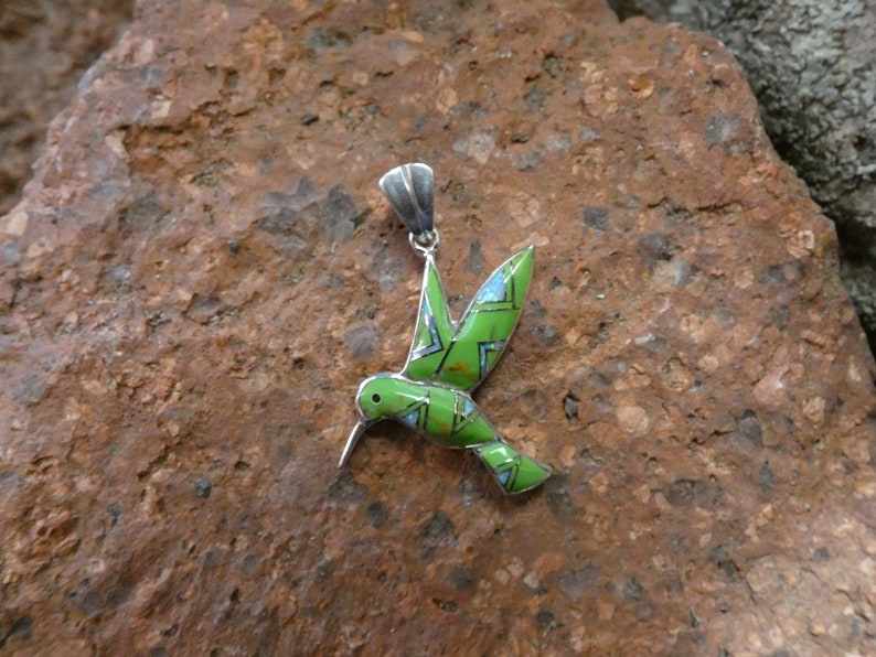 May include: A green and blue hummingbird pendant made of sterling silver. The hummingbird is in flight with its wings spread out. The pendant is a beautiful piece of jewelry that would make a great gift.