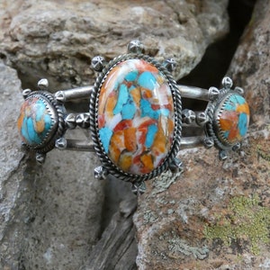 May include: A silver bracelet featuring three turquoise and orange stones set in a silver bezel. The stones are surrounded by a silver rope design.