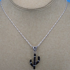 May include: A silver necklace with a cactus pendant. The pendant is black with white and silver accents. The necklace is displayed on a neutral-colored surface.