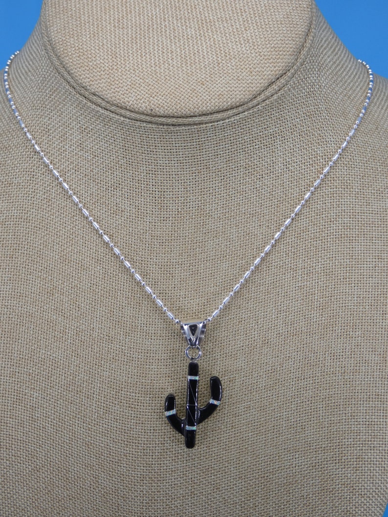 May include: A silver necklace with a cactus pendant. The pendant is black with white and silver accents. The necklace is displayed on a neutral-colored surface.