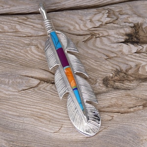 May include: A silver feather pendant with turquoise, orange, and purple inlay. The pendant is on a wooden surface.