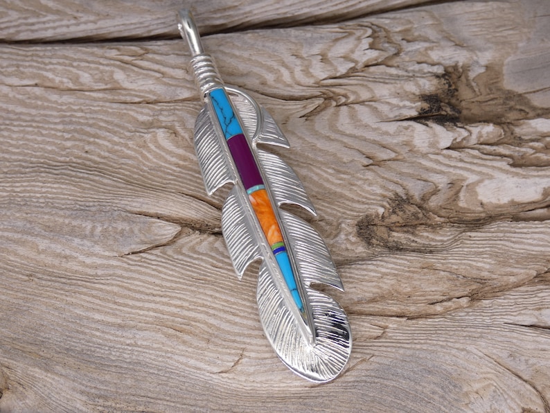 May include: A silver feather pendant with turquoise, orange, and purple inlay. The pendant is on a wooden surface.
