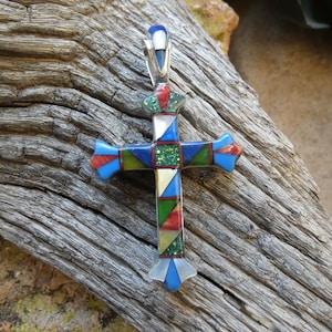 May include: A colorful, geometric cross pendant made of inlaid stones. The cross is made of silver and features a variety of colors, including blue, green, red, and white.