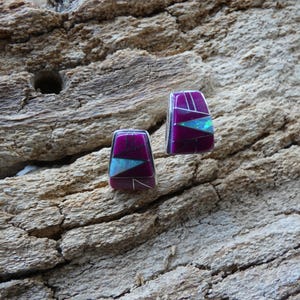 Puede incluir: Un par de pendientes de botón rectangulares con un diseño geométrico. Los pendientes presentan un fondo morado oscuro con acentos turquesa y blancos. Se encuentran sobre una superficie de madera marrón claro texturizada.