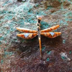 May include: A silver dragonfly pendant with orange and white inlaid wings and a central body. The wings feature a geometric pattern with a small, light blue inlay. The pendant is displayed against a textured, green and brown surface.