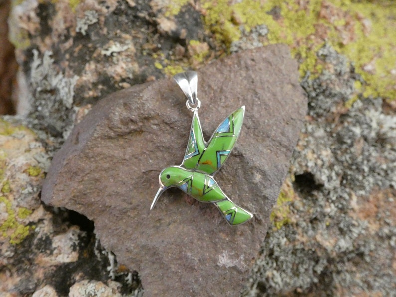 May include: A green and blue hummingbird pendant made of sterling silver with turquoise inlay. The pendant is in the shape of a hummingbird in flight.