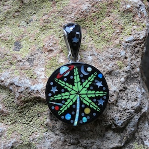 Puede incluir: Un colgante redondo de plata con fondo negro y un diseño de hoja de marihuana verde. El colgante está decorado con piedras de colores y estrellas.