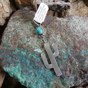 Puede incluir: Un colgante plateado con diseño de cactus y una piedra turquesa. El colgante hecho a mano descansa sobre una piedra texturizada y multicolor. La etiqueta dice "AMAL SPENCER NAVAJO HANDMADE SS KINGMA TURQ PENDANT".