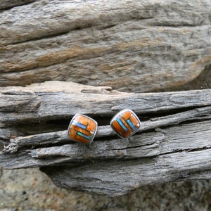 May include: A pair of square stud earrings featuring a mosaic inlay of orange spiny oyster shell and blue turquoise segments set within a silver frame. The earrings have a smooth, rounded square shape with a polished metal border.