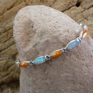 Könnte beinhalten: Ein silberfarbenes Gliederarmband mit abwechselnd angeordneten ovalen Steinen. Das Armband zeigt ein wiederkehrendes Muster aus schillernden blauen Opalen und glatten, einfarbigen orangefarbenen Steinen in polierten Metallfassungen.