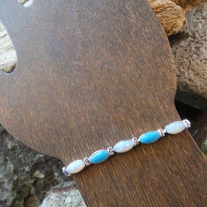 May include: A silver bracelet featuring oval-shaped turquoise and white opal stones. The bracelet is displayed on a wooden hand-shaped form, showcasing the alternating colors and the delicate design of the jewelry.