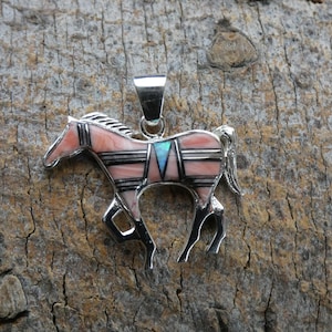 Op de afbeelding: Een zilveren paardenhanger met een roze en zwart ingelegd ontwerp. Het paard heeft een turquoise accent en staat tegen een getextureerde, bruine achtergrond. De hanger is een gedetailleerd sieraad.