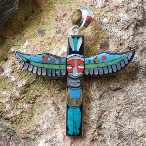 Puede incluir: Un colgante de plata con un diseño de tótem nativo americano colorido con un pájaro en la parte superior. El tótem está hecho de varias piedras y se coloca sobre un fondo rocoso.