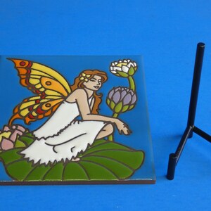 May include: A ceramic tile with a colorful illustration of a fairy sitting on a lily pad with wings spread. The fairy is wearing a white dress and has brown hair. The tile is on a black metal stand.