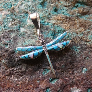 May include: A silver dragonfly pendant with turquoise and white inlay. The dragonfly's wings are a vibrant turquoise, with white accents. The pendant has a silver bail for hanging.