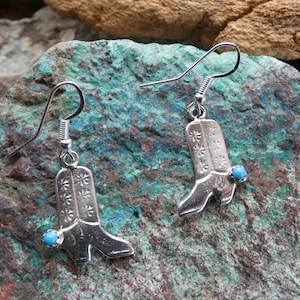 May include: A pair of silver-toned cowboy boot earrings. Each boot has a floral design and a small, round, turquoise-colored stone. The earrings hang from a simple hook.
