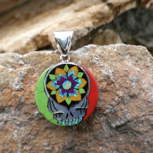 May include: A round silver pendant with a green, black, and red background. A colorful flower with a purple center is in the center of the pendant. The pendant is on a rock.
