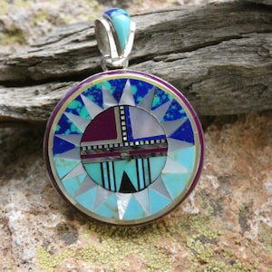 Puede incluir: Colgante circular con un diseño incrustado detallado. El colgante presenta una fianza plateada y un diseño central con turquesa, lapislázuli, nácar y otras piedras de colores. El anillo exterior es de color púrpura oscuro.