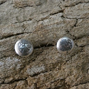 May include: Two silver stud earrings with a Kokopelli design. The earrings are round and feature a detailed, stylized image of the Kokopelli figure. The earrings are set against a textured, brown background.