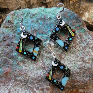 May include: A jewelry set featuring a pendant and matching earrings. The black, diamond-shaped pieces are adorned with celestial designs, including moons, stars, and colorful accents. The earrings have silver hooks.
