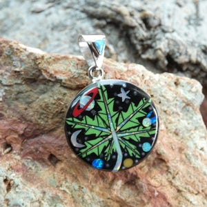 Puede incluir: Un colgante de plata con una hoja verde sobre fondo negro y detalles celestes coloridos, incluyendo un planeta, estrellas y una luna creciente.