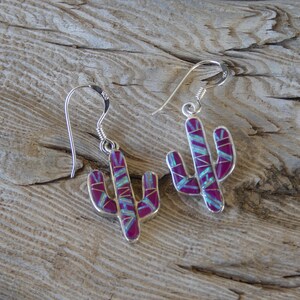 May include: A pair of silver cactus earrings with a magenta and turquoise inlay. The earrings are shaped like cacti with detailed segments and a hook closure. The earrings are displayed on a weathered wooden surface.