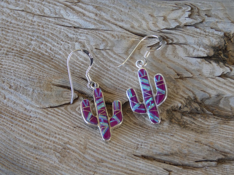 May include: A pair of silver cactus earrings with a magenta and turquoise inlay. The earrings are shaped like cacti with detailed segments and a hook closure. The earrings are displayed on a weathered wooden surface.