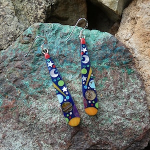 May include: A pair of dangle earrings with a celestial theme. The earrings have a dark blue background with white stars, crescent moons, and colorful circles. The bottom is yellow and the top is red.