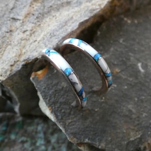 May include: A pair of silver-toned hoop earrings with inlaid white, blue, and black stones. The earrings are curved and rest on a textured gray rock surface. The stones are arranged in a repeating pattern.