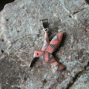 May include: A hummingbird pendant featuring inlaid segments of pink stone and iridescent blue accents set within a silver frame. The bird is depicted in flight with wings extended and a long, thin beak, suspended from a small silver bail at the top.